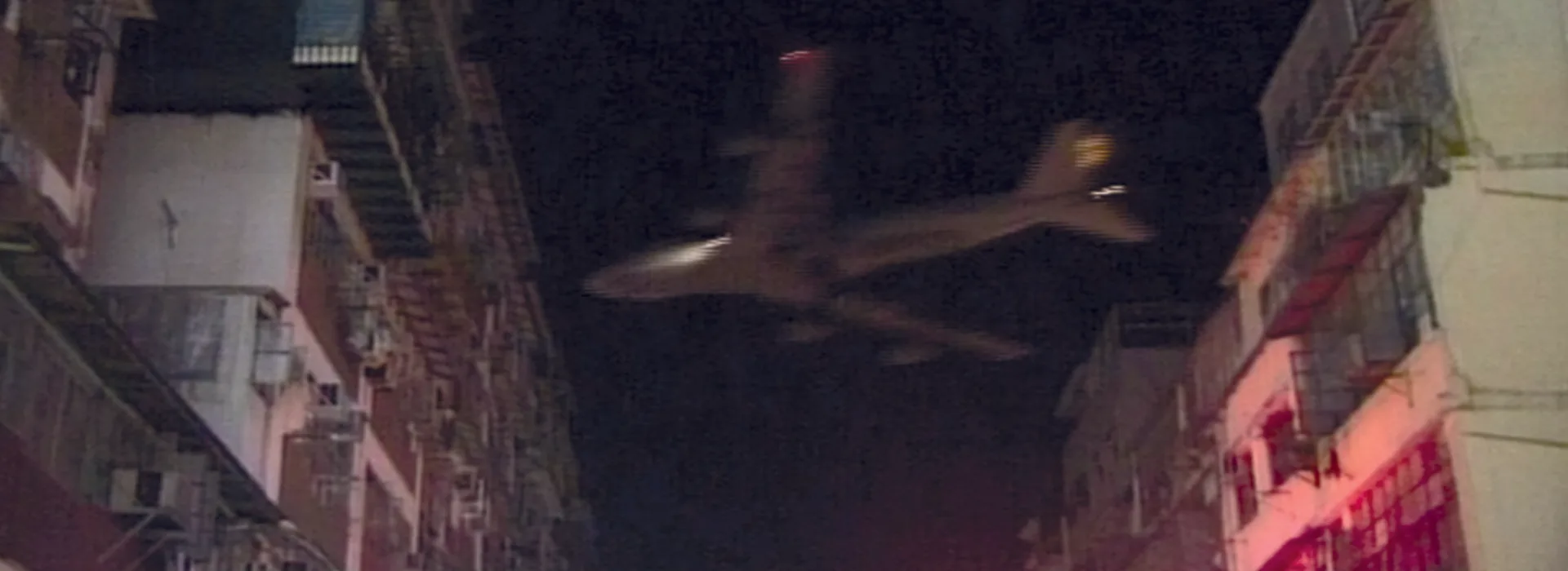Nächtliche Strassenschlucht in Hongkong mit Neonreklamen und einem tief fliegenden Flugzeug