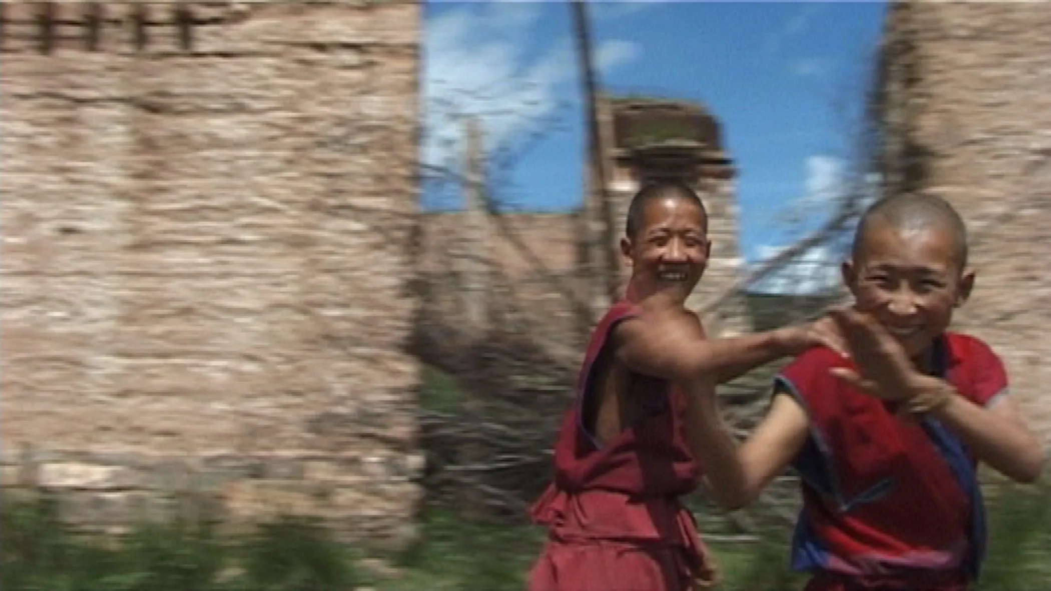Vignette du film Angry Monk (2005) : Deux jeunes moines tibétains courent en riant devant d’anciennes ruines de pierre et regardent la caméra.