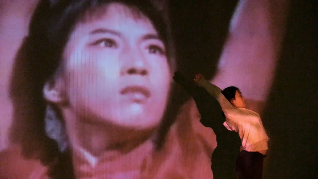 A dancer stands before a large projection of a woman’s face.